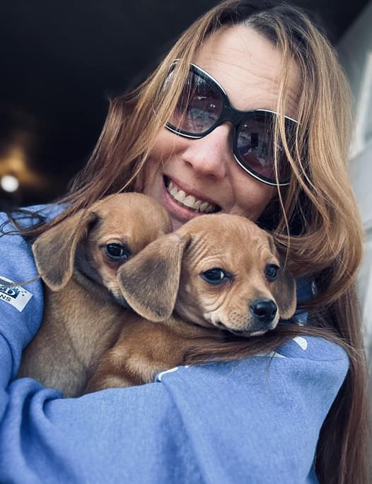woman facing forward smiling holding two puppies
